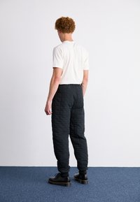 Pantalon matelassé noir avec taille élastique, associé à une chemise blanche à manches courtes. Le modèle se tient de dos, avec des chaussures noires visibles.
