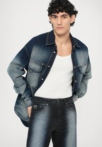 Denim srajca v gradientni modri barvi, z dolgimi rokavi in dvema žepoma na prsih. Oblečena čez belo majico brez rokavov in kombinirana s temnimi denim jeans hlačami z izbledelimi detajli.