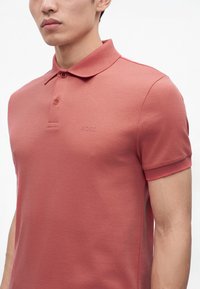 Homme portant un polo cintré à manches courtes de couleur rose poudré avec deux boutons et un logo discret "BOSS" sur la poitrine.