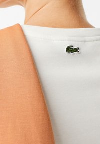 Nära bild av en persons nacke och övre rygg som bär en vit skjorta med en grön krokodil-logotyp och ett orange plagg hängande över axeln.