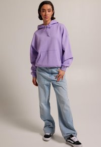 Sudadera oversized de lavanda con bolsillo frontal, combinada con jeans de pierna ancha de color azul claro y zapatillas negras con suelas blancas. Fondo simple.