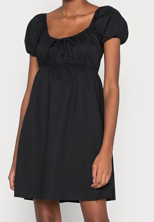 Frau trägt ein kurzes schwarzes Kleid mit Puffärmeln und einem quadratischen Ausschnitt, der in der Mitte mit einer kleinen Schleife gebunden ist.