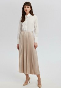 Valkoinen kauluspaita yhdistettynä pleated-beige-hameeseen. Hameessa on lentävä rakenne, joka ulottuu polven alapuolelle, ja se on täydentynyt nuden värisillä korkokengillä.