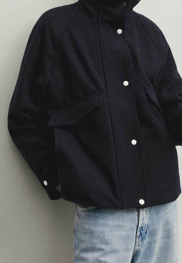 Veste bleu marine à boutons avec un col haut, deux poches avant et des manches raglan, fabriquée dans un tissu doux et texturé.