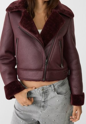 Kobieta w krótkiej bordowej kurtce z owczej skóry z kieszeniami na suwak i jasnoszarych dżinsach z metalowymi ćwiekami, z ręką schowaną w przedniej kieszeni.