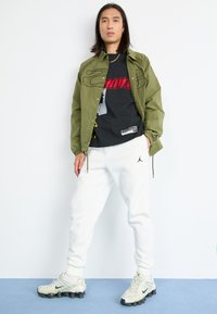 Olijfgroene jas, zwart graphic t-shirt, witte joggers en lichtgrijze sneakers met zwarte accenten. Accessoires zijn een gouden ketting.