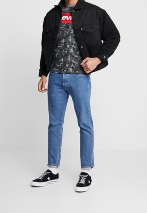 Černá bunda s ovčí vlnou, šedé tričko s kamuflážním vzorem a červeným logem "Levi's", světle modré džíny a černé tenisky s bílými detaily.