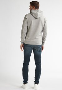 Hombre de pie de espaldas, vistiendo una sudadera gris claro, jeans azul oscuro y zapatillas blancas, contra un fondo plano.