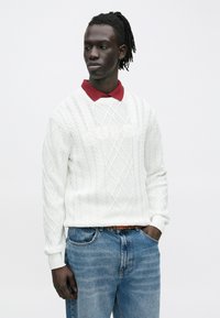 Mężczyzna z dredami ubrany w biały sweter w warkocze na czerwonej koszulce polo i niebieskich dżinsach, stojący z rękami w kieszeniach, patrzący w bok.