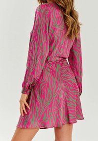 Mujer vistiendo un vestido de zebra rosa y verde de manga larga con cintura ajustada, mostrado desde atrás con el cabello suelto y ondulado.