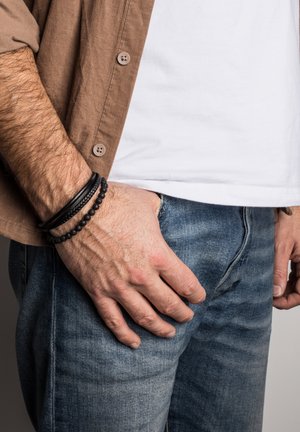 Black and textured bracelets adorn a wrist, alongside a light brown corduroy shirt and blue denim jeans, with a white T-shirt underneath.