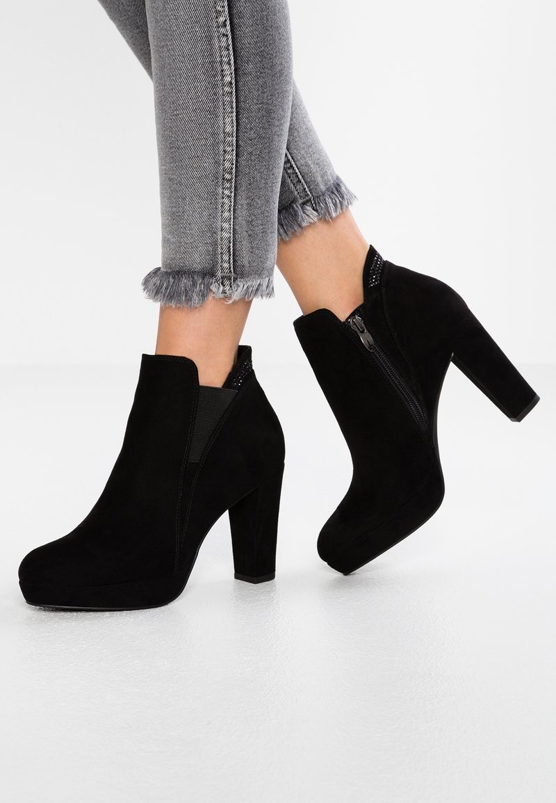 Bottines en daim noir à talons blocs portées avec un jean gris à ourlet effiloché, un pied légèrement levé pour montrer le détail de la fermeture éclair latérale.