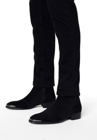 Black suede ankle boots with a low block heel, featuring a metallic toe cap and a side zipper, paired with black slim-fit pants.