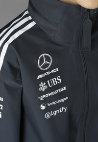 Crna jakna s bijelim prugama, logotipima i brendiranjem. Materijal izgleda glatko s patentnim zatvaračem i dizajnom visokog ovratnika.