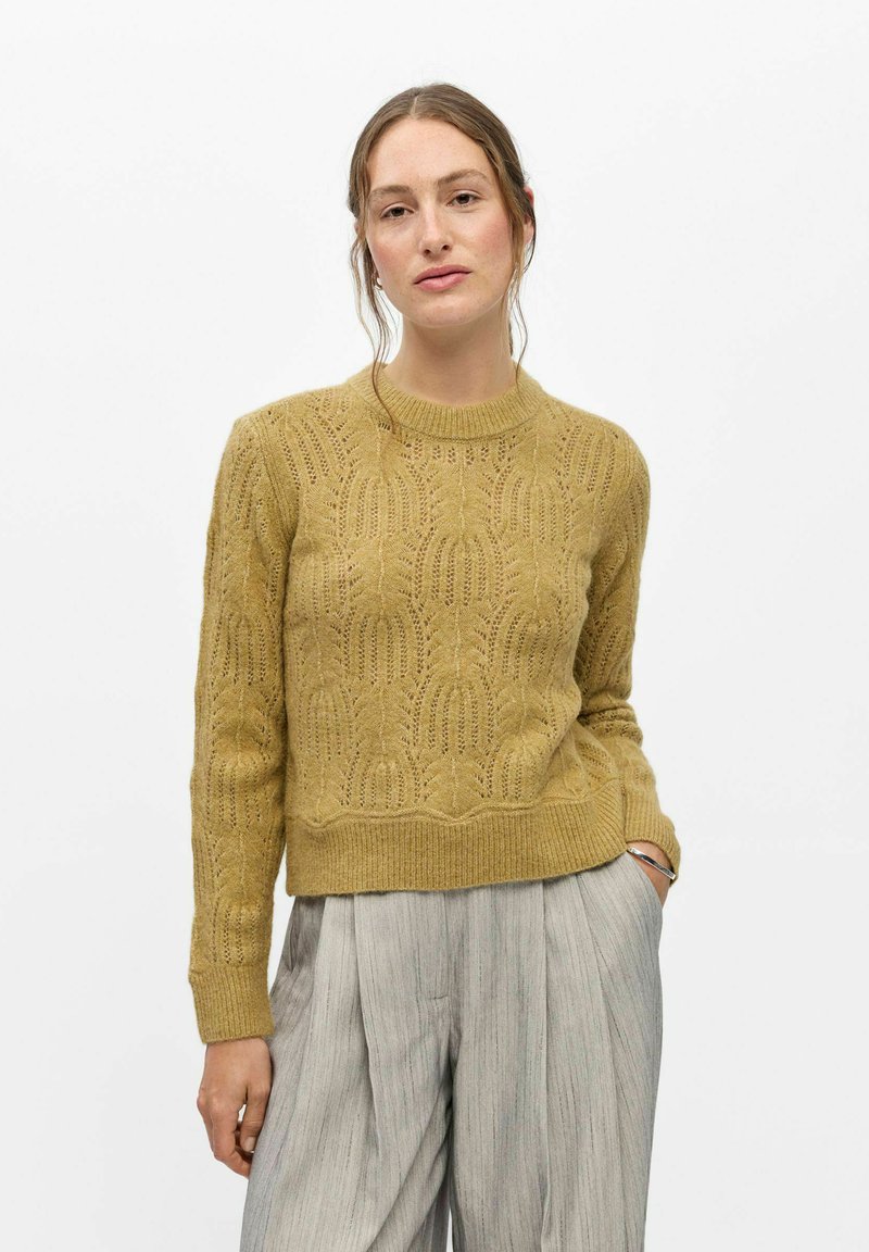 Femme aux cheveux tirés en arrière porte un pull en tricot jaune moutarde et un pantalon large plissé gris clair, debout avec une main dans la poche.