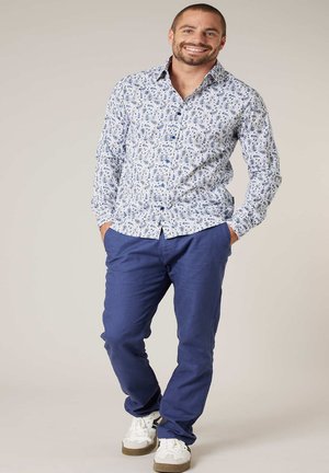 Glimlachende man met blauw patroon lange mouwen shirt, blauwe broek en witte sneakers staat met handen in zakken tegen een effen achtergrond.