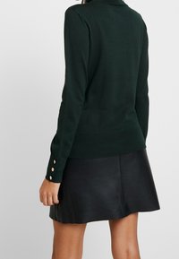Dunkelgrüner Strickpullover mit langen Ärmeln und geripptem Saum. Hat goldene Knopfdetails an den Bündchen. Wird mit einem schwarzen Bleistiftrock aus Leder getragen.