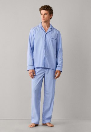 Jeune homme debout pieds nus portant un pyjama bleu clair boutonné à manches longues avec bord foncé au col et aux poignets, fond gris neutre.
