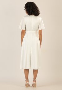 Weißes Satin-Kleid mit kurzen Ärmeln, tailliertem Schnitt und fließendem Rock. Der Rücken hat einen geraden Ausschnitt und eine gebundene Gürtel-Detail.