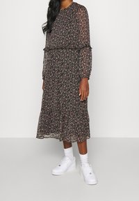 Kobieta w czarnej midi sukience w mały różowy kwiatowy wzór, z długimi przezroczystymi rękawami, białymi skarpetkami i białymi sneakersami na tle jednolitym.