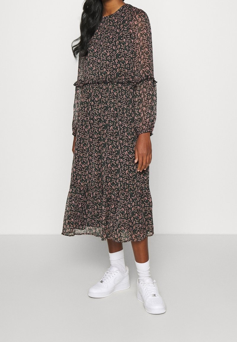 Kobieta w czarnej midi sukience w mały różowy kwiatowy wzór, z długimi przezroczystymi rękawami, białymi skarpetkami i białymi sneakersami na tle jednolitym.