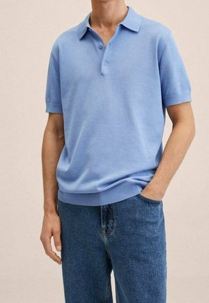 Henkilö pukeutuneena vaaleansiniseen lyhythihaiseen poolopaitaan, joka on osittain tungettu sinisiin farkkuihin, toinen käsi taskussa.