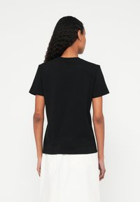 Donna con capelli ricci che indossa una maglietta nera semplice a maniche corte e una gonna bianca, in piedi di spalle contro uno sfondo bianco.