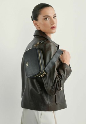 Kobieta z ciemnymi włosami związanymi w koka, ubrana w czarną skórzaną kurtkę i białe spodnie, trzymająca na ramieniu czarną torebkę na ramię z złotymi zamkami błyskawicznymi.