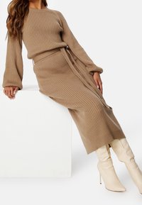 Femme portant une robe en maille côtelée beige à manches longues avec une ceinture nouée, associée à des bottes à talons hautes crème, appuyée sur un bloc blanc.