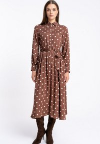 Brą dress w kolorze brązowym, z długim rękawem, białe kropki, kołnierzyk zapinany na guziki i pasek w talii; zwiewny materiał i długość do kolan, noszone z brązowymi butami.