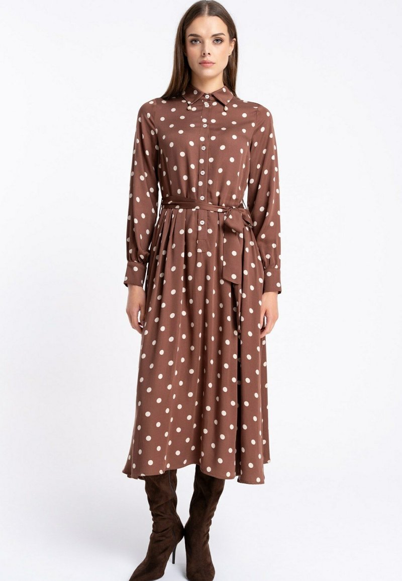 Brą dress w kolorze brązowym, z długim rękawem, białe kropki, kołnierzyk zapinany na guziki i pasek w talii; zwiewny materiał i długość do kolan, noszone z brązowymi butami.