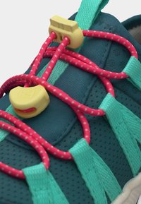 Blågrønn joggesko med rosa elastiske snører uten knyting, sikret med to gule snørelåser på perforert stoff overdel.