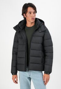 Mand iført en sort puffjakke med hætte over en mørkegrøn sweater og lyseblå jeans, stående foran en ensfarvet hvid baggrund.