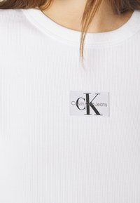 Bílý žebrovaný tričko s kulatým výstřihem. Na hrudi má obdélníkovou nášivku s nápisy "CK" a "Calvin Klein Jeans" v černém písmu.