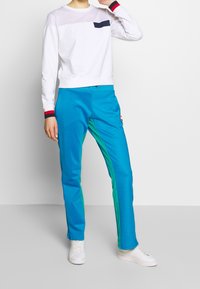 Biała bluza z siateczkowym panelem i granatową kieszenią, w zestawieniu z jasnoniebieskimi spodniami z zielonymi bocznymi paskami i naszywką z logo. Białe sneakersy.