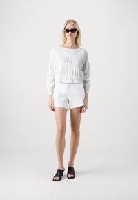 Top bianco a maniche lunghe con texture e pieghe, abbinato a pantaloni shorts in denim bianco sfilacciati e mule nere con punta aperta. Occhiali da sole scuri completano il look.