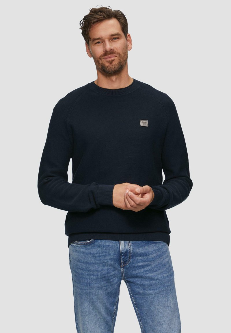 Marineblauwe sweatshirt met ronde hals, ribgebreide manchetten en onderkant, met een gestructureerde stof en een klein logopatch op de borst, gecombineerd met lichte jeans.