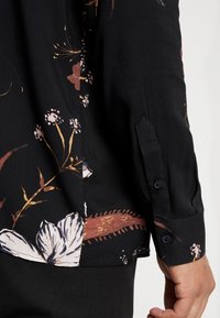 Chemise noire à motifs floraux, fabriquée dans un tissu doux, ornée de motifs vibrants de fleurs roses et crème, avec un détail de poignet boutonné.