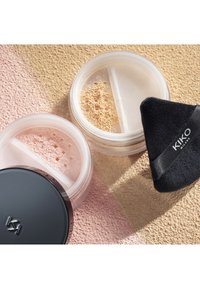 To gjennomsiktige krukker med løst pulver: en med rosa pulver, den andre med beige. En svart puff med logo ligger i nærheten på teksturert bakgrunn.