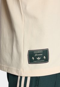 Krem boje pamučna majica s zelenom etiketom na kojoj piše "Juventus" i logotipom, smještena na donjoj strani. Glatka tekstura, regularni kroj.