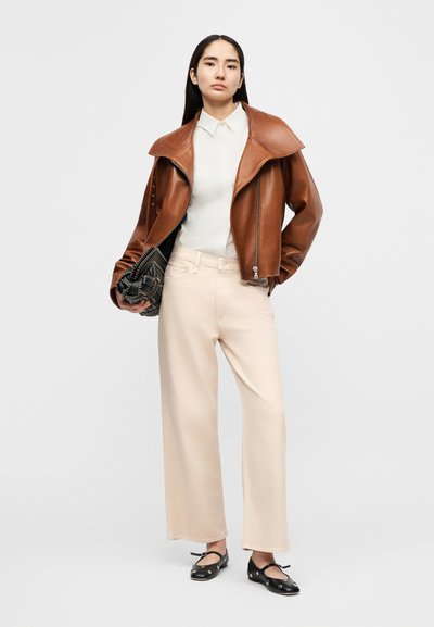 Γυναίκα με καφέ δερμάτινο μπουφάν πάνω από λευκό πουκάμισο, μπεζ παντελόνι με φαρδιές μπατζάκια, κρατώντας μια μαύρη τσάντα, όρθια με το ένα χέρι στην τσέπη.