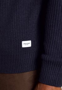 Närbild av mörkblå ribbad tröjärm och kropp med vit Jack & Jones-märkesetikett ovanför bruna byxor.