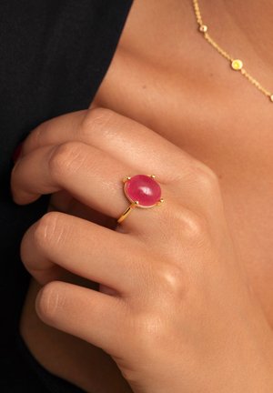 Anillo de oro con una piedra rosa redonda, superficie lisa y acabado pulido. Mano con un tono de piel natural sosteniendo el anillo.
