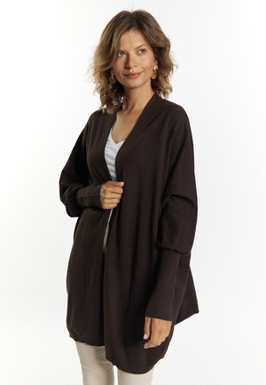 Brun, oversized cardigan med en blød tekstur, lange ærmer og åbent frontdesign. Bæres over en hvid top og beige bukser.