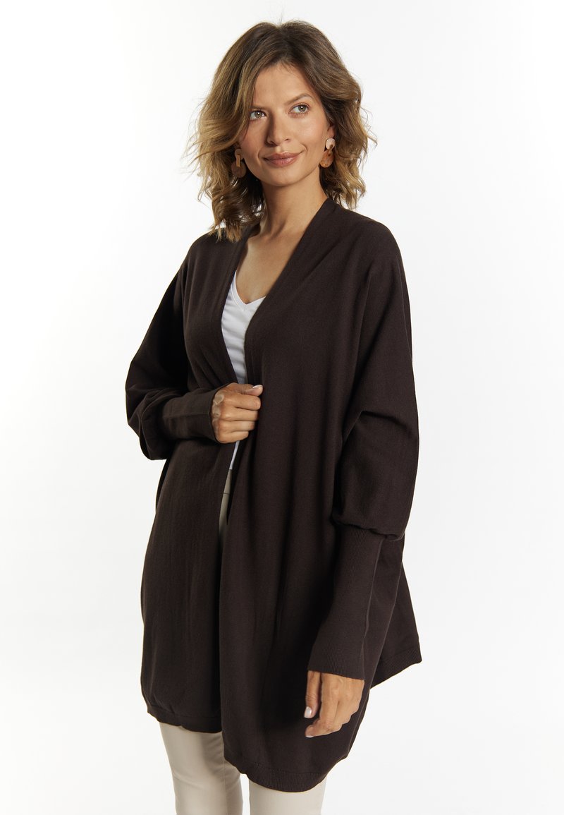 Brun, oversized cardigan med en blød tekstur, lange ærmer og åbent frontdesign. Bæres over en hvid top og beige bukser.