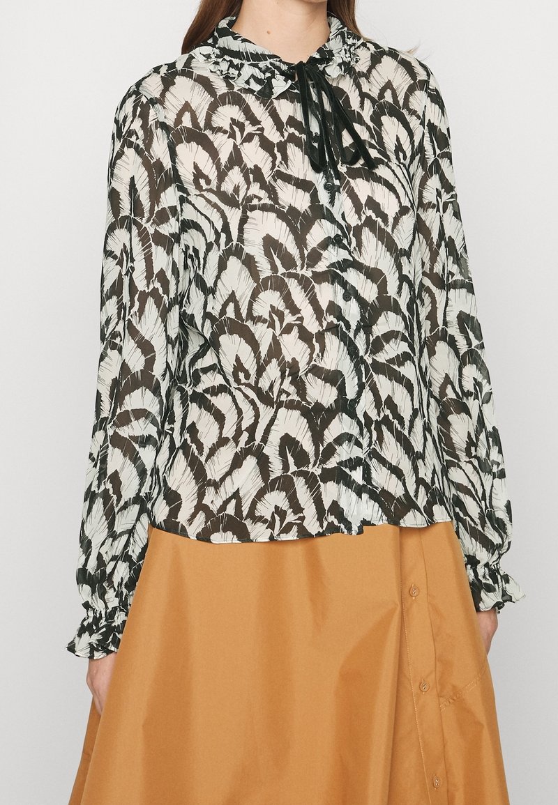 Blusa trasparente bianca e nera con motivo a foglia, polsini arricciati e lacci al collo, abbinata a una gonna arancione uniforme con bottoni laterali.
