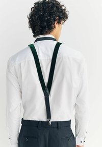 Green velvet suspenders attach to a black leather yoke, worn over a white shirt and black pants, with adjustable clips for secure fit.
