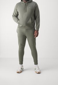 Felpa verde oliva e pantaloni jogger abbinati in morbido tessuto; la felpa ha una tasca frontale e un cordino. Le scarpe da ginnastica sono bianche con dettagli verdi.