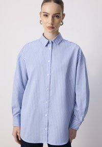 Giovane donna che indossa una camicia a righe verticali blu e bianche con maniche lunghe e orecchini a cerchio in argento su uno sfondo neutro.