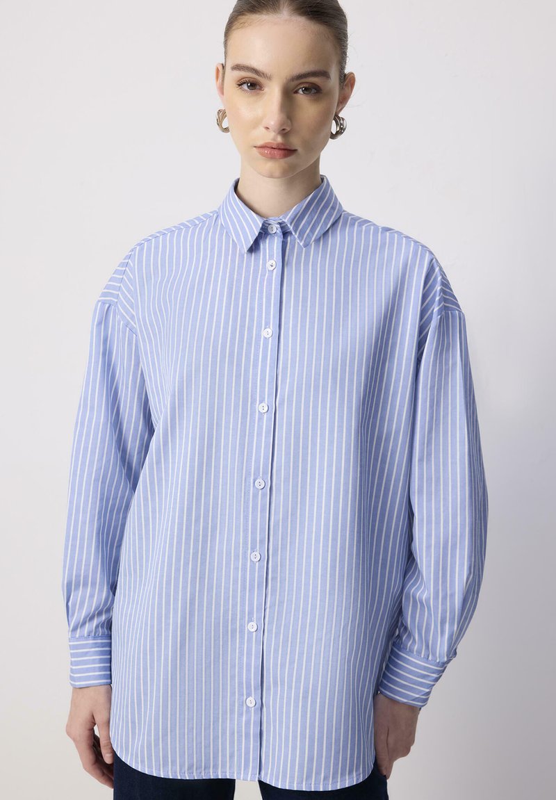 Giovane donna che indossa una camicia a righe verticali blu e bianche con maniche lunghe e orecchini a cerchio in argento su uno sfondo neutro.